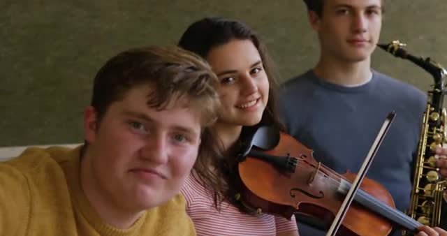 Three teenagers in a music rehearsal setting, with one girl holding a violin and a user saxophone. They smile at the camera, creating a friendly and inclusive atmosphere. This can be great for educational websites, music school brochures, and articles promoting youth musical programs. Highlights teamwork, creativity, and learning.