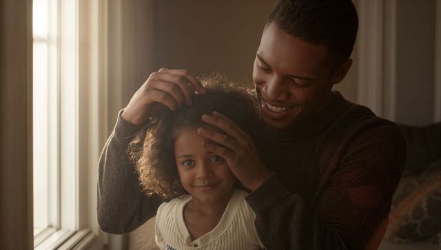 Sunlit father styling daughter's curly hair by window, cozy warm family bonding moment