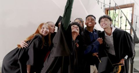Multicultural graduates celebrating and taking selfie in foyer wearing caps and gowns