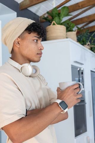 African american man standing in kitchen holding ribbed mug wearing beanie and headphones