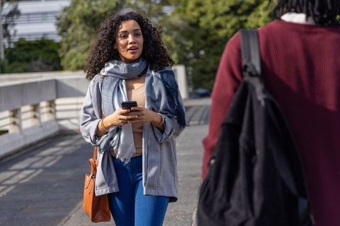 Young woman walking holding smartphone and carrying brown tote bag on urban walkway