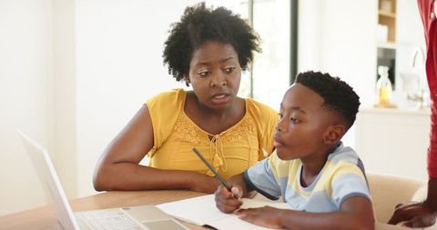 African American mother helping son with homework at kitchen table using laptop and notebook