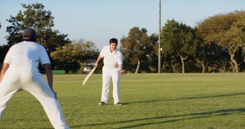 Indian batter swinging wooden bat while teammate fielding on sunny park cricket ground