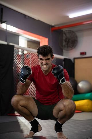 Aggressive mma fighter crouching on mats in gym wearing red shirt and black gloves expressing fierce