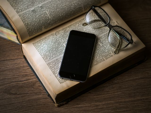 Smartphone and glasses placed on open vintage book, all on wooden desk. Ideal for themes of modern learning, combining technology with traditional methods and academic pursuits. Could be used for educational materials, technology blending with literature, or reading promotions.