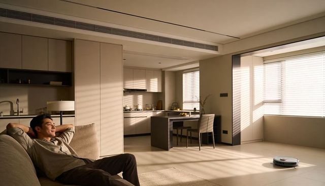 Sunlit Minimalist Open-Plan Living Room with Relaxed Asian Man on Sofa and Robot Vacuum