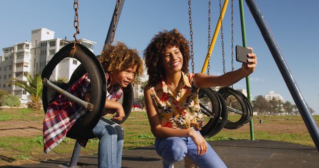 Happy biracial mother taking selfie with son on swing at sunny playground. Communication, motherhood, childhood, togetherness, summer, vacations, fun and free time, unaltered.