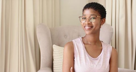 Smiling african woman sitting in beige wing chair, wearing glasses and hoop earrings