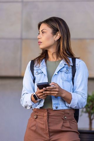 Young woman checking smartphone outside wearing denim jacket and backpack, urban lifestyle