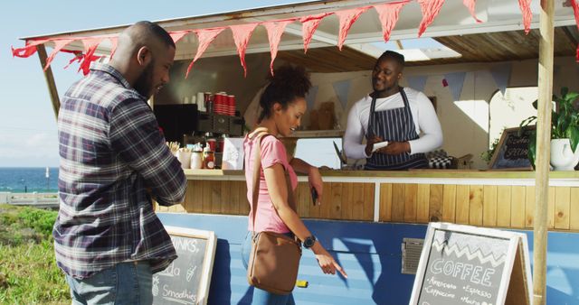African American couple ordering coffee at a food truck located near a beach. The barista in an apron is preparing their order while the vibrant atmosphere enhances the urban coastal vibe. Ideal for use in promotional materials for street food vendors, coffee stands, food and beverage advertisements, or travel blogs showcasing trendy, outdoor eating spots.