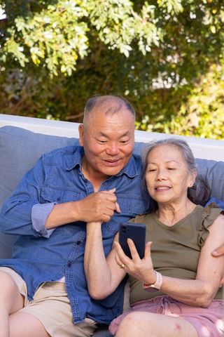 Senior asian couple sharing smartphone on sunlit backyard sofa, holding hands