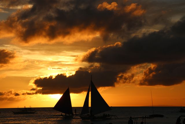 Silhouettes of sailboats gliding on the ocean at sunset with dramatic clouds in the sky. This tranquil scene is ideal for themes related to travel, relaxation, serenity, adventure, and seaside vacations. Suitable for travel brochures, websites, posters, and social media about sailing, exploration, and coastal life.