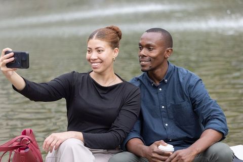 Smiling couple taking lakeside selfie with coffee and red handbag, casual outdoor date