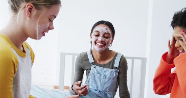 Three diverse young women are enjoying a relaxing day at home with face masks. They are laughing and bonding while applying skincare treatments in a cozy atmosphere. Perfect for concepts related to friendship, relaxation, skincare routines, self-care, and youthful happiness.