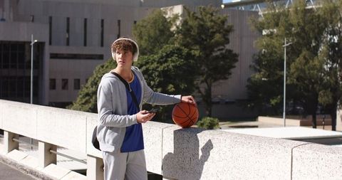 Teen male listening to music holding basketball on urban campus overpass with backpack