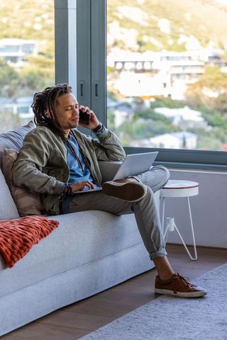 African american man working remotely on laptop while talking on phone in sunlit apartment