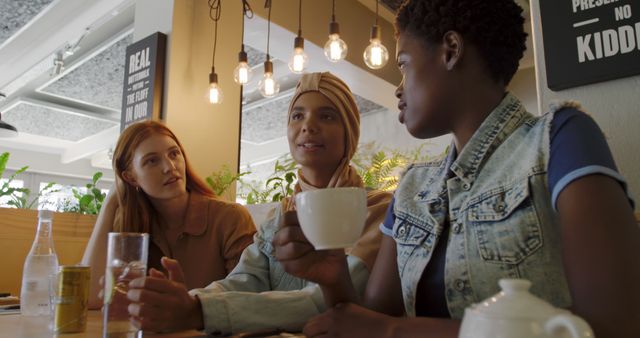 Three diverse friends engaged in conversation at a cafe while enjoying beverages. Seen with modern casual attire and illuminated by hanging lightbulbs, they embody a relaxed and inclusive atmosphere. Perfect for depicting social gatherings, inclusivity, and modern lifestyles in marketing, social media posts, and advertisements.