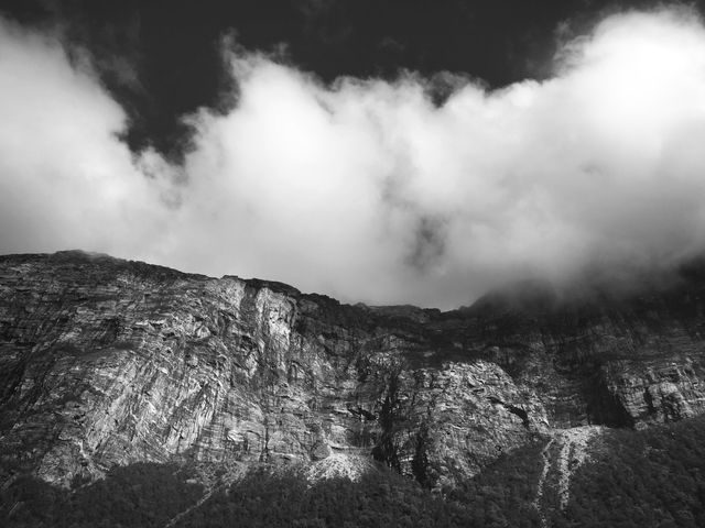 A dramatic black and white landscape capturing a rugged mountain partially enveloped by clouds. Useful for backgrounds, travel advertisements, nature documentaries, or prints for home décor. Emphasizes tranquility and the grandeur of rugged natural beauty.