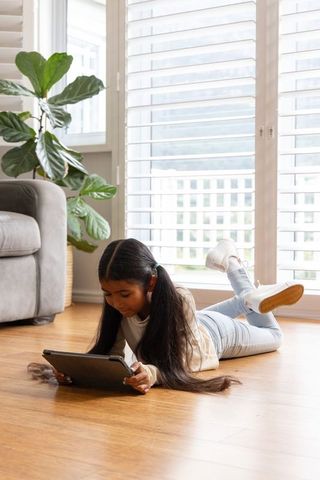 Asian girl using tablet while lying on hardwood floor in sunlit modern living room