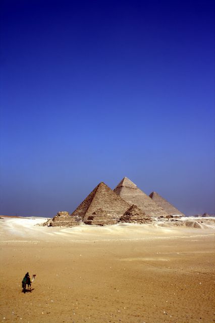 Solitary traveler walking across a desert towards the ancient Pyramids of Giza in Egypt. The scene showcases the vastness of the desert and the grandeur of the pyramids against a clear blue sky. Suitable for travel promotions, historical articles, adventure blogs, or educational materials about ancient civilizations and archaeology.