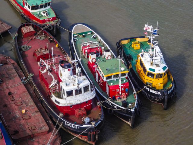 Tugboats with various colors, including red, green, and yellow, are moored side by side in a harbor. This aerial perspective captures the intricate details and vibrant hues of the ships. Useful for maritime industry, transportation hubs, or nautical themes. It can also be used in content related to commercial shipping, port operations, or seaside tourism.