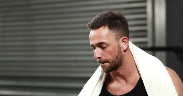 A middle-aged Caucasian man appears exhausted after a workout, with a towel around his neck and copy space. His expression and the gym setting suggest he's taking a moment to rest and recover from intense physical exercise.