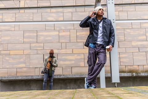 Diverse young men in streetwear leaning on column with coffee and headphones in city plaza
