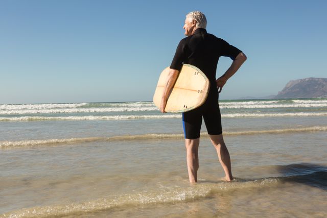 Senior man standing at the edge of the ocean holding a surfboard, ready to surf. Ideal for use in advertisements promoting active lifestyles, retirement activities, fitness, and outdoor adventures. Suitable for articles or blogs about surfing, senior fitness, and beach vacations.