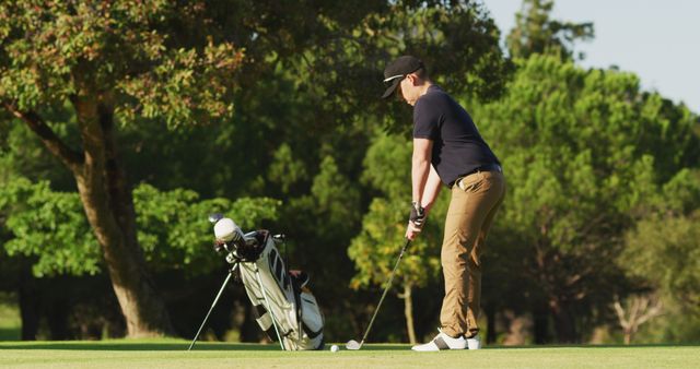 Golfer is preparing to take a swing on a well-maintained golf course during a sunny day. Golf bag and clubs are standing nearby. Image is ideal for illustrating themes related to sports, outdoor activities, leisure, golf training, golf equipment, and healthy lifestyle.