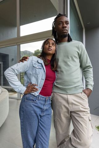 Young diverse couple standing and gazing through modern home floor-to-ceiling glass doors
