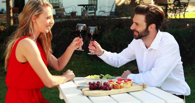 A Caucasian man and woman enjoy a romantic outdoor meal, toasting with glasses of wine, with copy space. Their cheerful expressions and elegant attire suggest a special occasion or celebration.