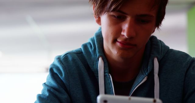 Teenage boy wearing blue hoodie concentrating on smartphone usage indoors. Useful for illustrating adolescent lifestyle, mobile technology, connectivity in modern youth, or social interaction scenes.
