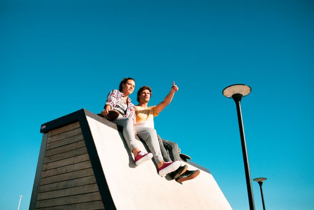 Two male and female friends sitting on wall and talking at skatepark. hanging out at an urban skatepark in summer.