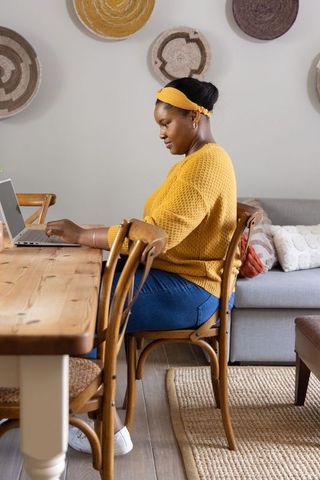 African american woman working from home in cozy mustard sweater at wooden dining table