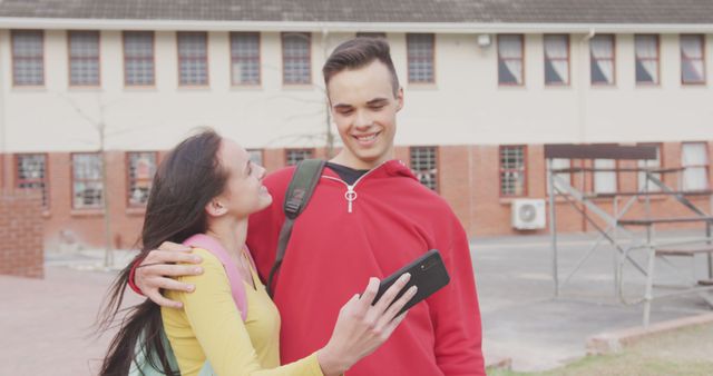 Happy caucasian couple taking selfie using smartphone by school building. Secondary school, education, technology, photography, relationship and teenage hood concept, unaltered.