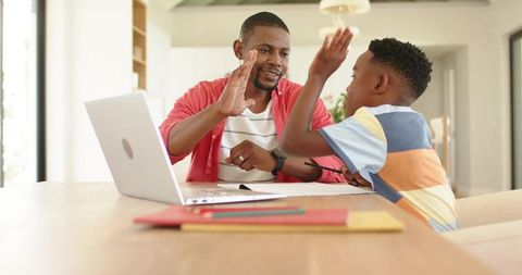 African american father and son high-fiving while homeschooling at kitchen table, celebrating learni