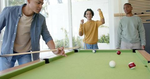Young men playing pool on green felt table in bright modern lounge celebrating casual shot