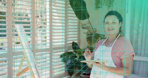 Mature woman painting on canvas in sunlit studio wearing white apron teal grid overlay