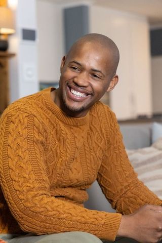 African american man smiling while sitting on sofa wearing mustard cable-knit sweater