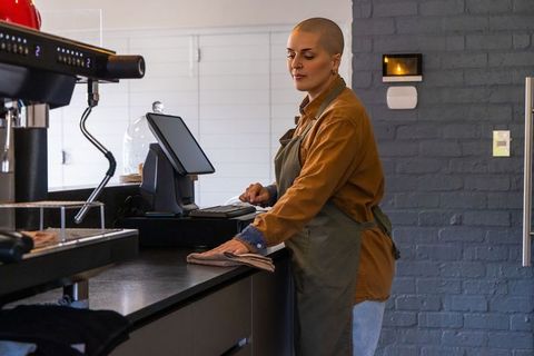 Barista wiping counter while using pos touchscreen in modern coffee shop interior