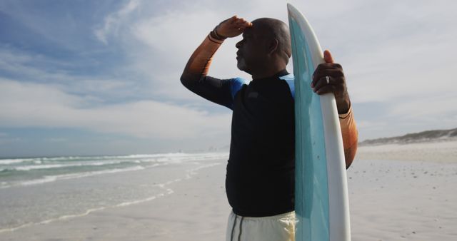 An African American man is holding a surfboard on a beach and scanning the horizon under a partly cloudy sky. Ideal for promoting outdoor activities, surfing sports, active lifestyles, travel destinations, vacation experiences, or fitness campaigns.