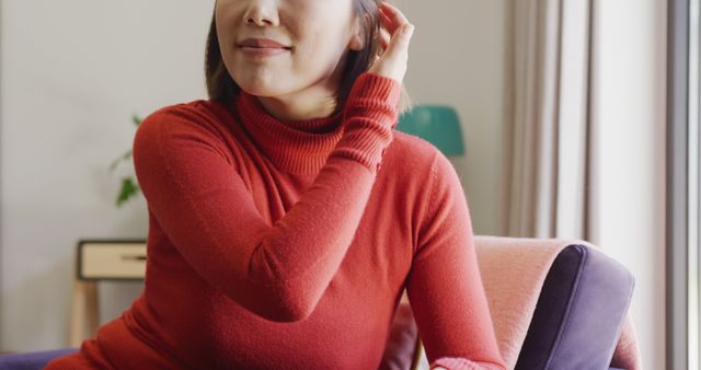 Asian woman in red sweater casually touching her hair while sitting on a sofa at home. Ideal for lifestyle blogs, home living magazines, fashion editorials focused on casual wear, or advertisements promoting relaxation and comfort in home settings.