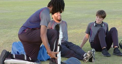 Diverse cricket teammates laughing and unpacking pads during grass field break