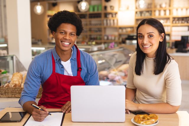 Two young cafe owners, one African American and one biracial, are sitting at a table in their cafe, smiling and working on a laptop. The cafe interior is modern and well-lit, with various food items visible in the background. This image can be used for promoting small businesses, teamwork, entrepreneurship, and the food industry.