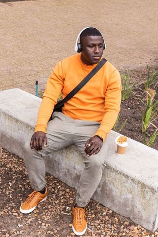 African american man sitting in orange sweater with headphones and coffee on urban bench
