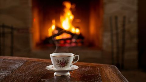 Steaming porcelain teacup with rose design resting on polished wood near roaring fireplace