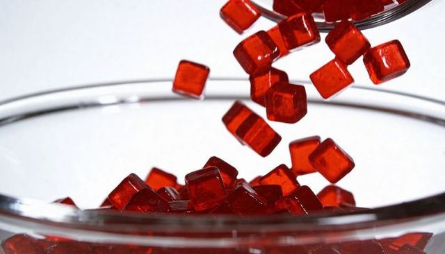 Pouring red gummy cube candies into glass bowl with metal scoop, glossy closeup macro