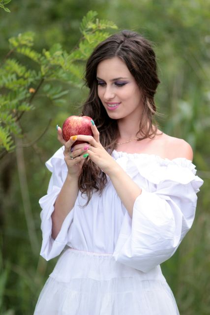 Woman in a white dress holding an apple, standing in a green garden. Ideal for concepts of health, nature, beauty, and lifestyle. Can be used for advertising healthy living, nature retreats, or natural beauty products.