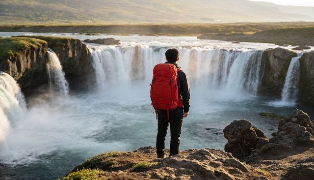 Hiker standing on cliff edge facing dramatic waterfall, carrying red backpack at sunrise