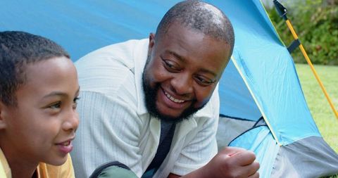 Father and son camping together, smiling inside blue tent, bonding outdoors on grassy lawn